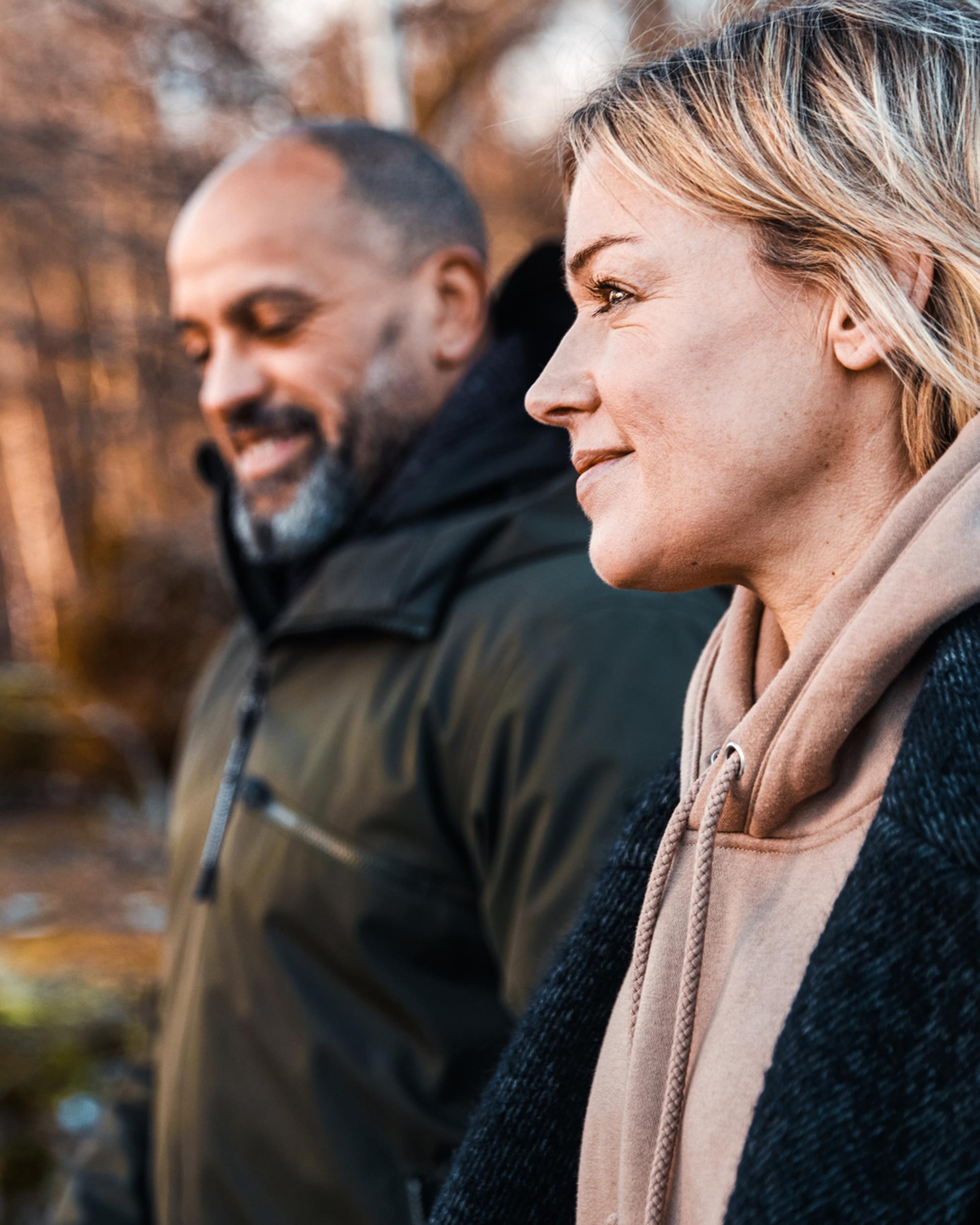 Två personer promenerar utomhus i en höstig miljö. Kvinnan i förgrunden ser nöjd ut medan hon bär en beige hoodie och en mörk kappa. Mannen i bakgrunden bär en mörk jacka