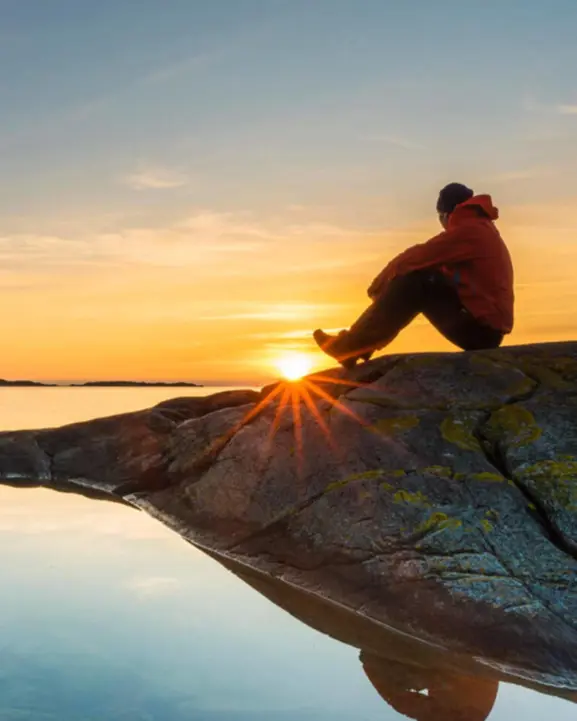 Person sitter på klippa och ser solnedgången vid vattnet, medklar himmel och reflektioner i vattnet i Sverige