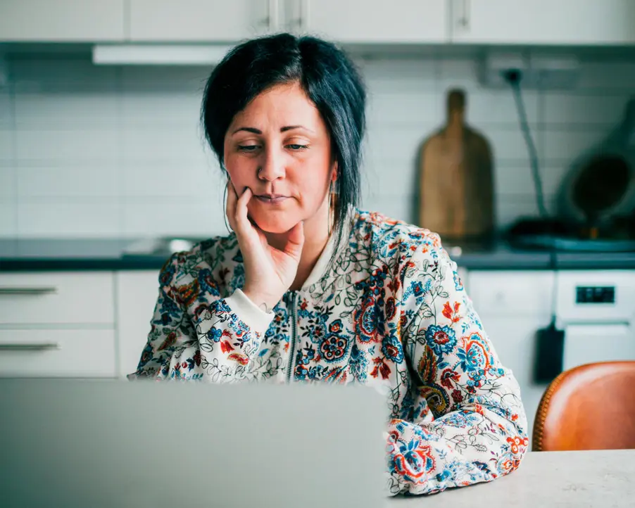 Kvinna sitter i kök med dator och mobil.
