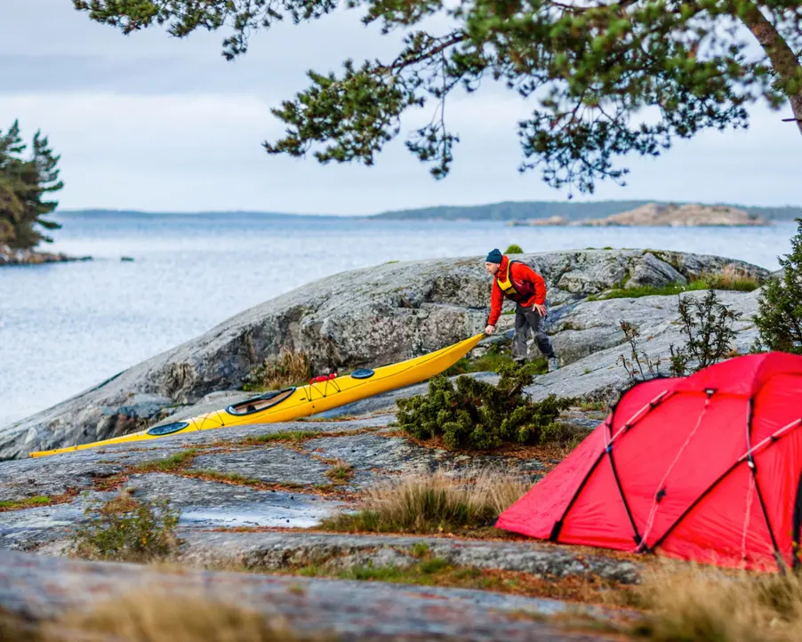 Person som drar en gul kajak på klipporna vid havet med ett rött tält i förgrunden, omgiven av svensk skärgårdsnatur