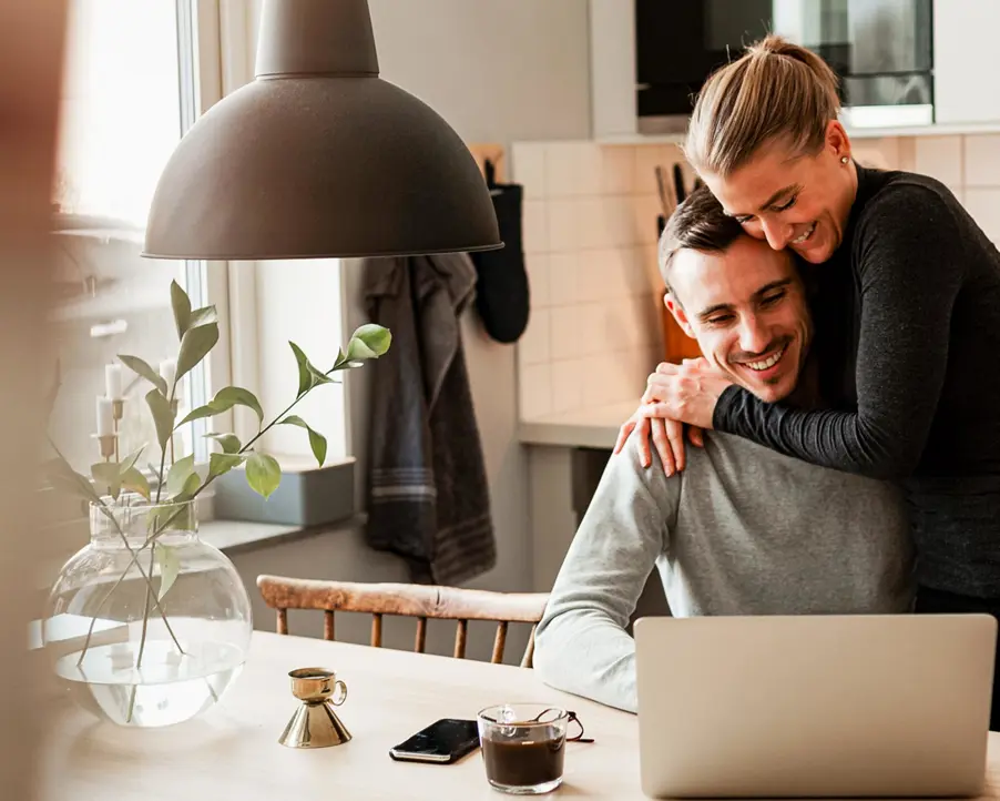 Par som arbetar på en bärbar dator vid köksbordet, leende och omfamnande, med en stilren inredning och en växt i bakgrunden