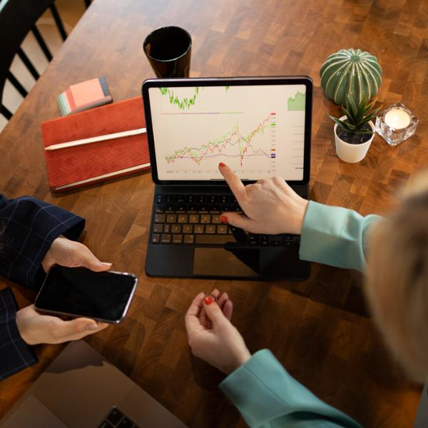 Två personer diskuterar aktiediagram på en bärbar dator vid ett träbord, med en smartphone och anteckningsböcker i närheten