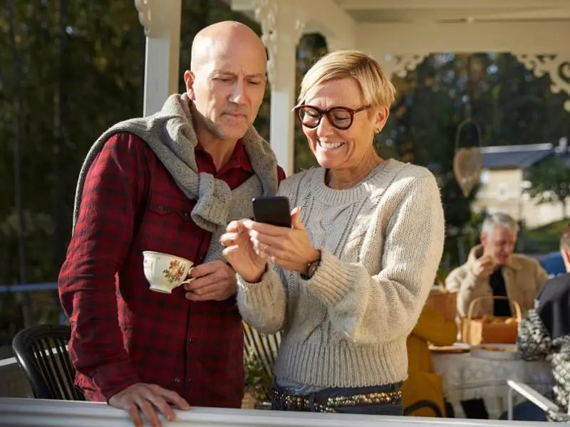 Äldre par på veranda med en tekopp, tittar på en mobiltelefon och ler. Solig utomhusmiljö