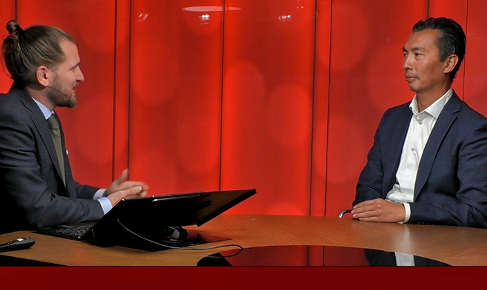 Två män i formell klädsel sitter mittemot varandra vid ett bord, har en diskussion framför en röd bakgrund