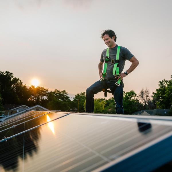 Person i s&#xE4;kerhetssele st&#xE5;r p&#xE5; ett tak med solpaneler i solnedg&#xE5;ngen. H&#xE5;llbar energiinstallation