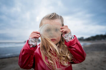 Flicka i röd jacka håller upp genomskinligt föremål vid havet