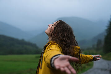 Kvinna i gul regnjacka med utsträckta armar i dimmigt bergslandskap