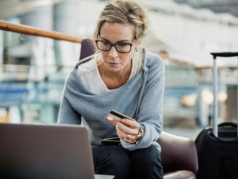 Kvinna med glasögon handlar online med kreditkort och laptop på en flygplats, nära en resväska