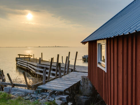 Röd stuga vid havet i solnedgång, med en träbrygga i förgrunden