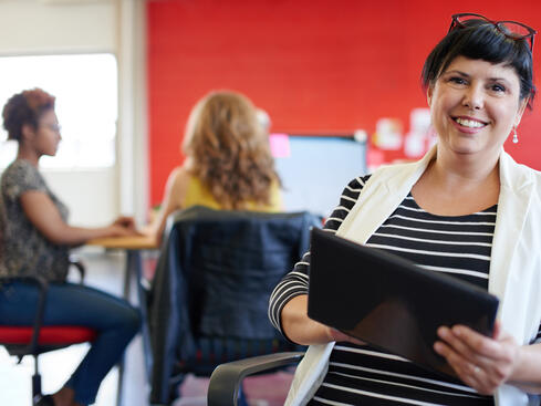 Kvinna i kontorsmiljö med surfplatta, leende, med två kollegor i bakgrunden