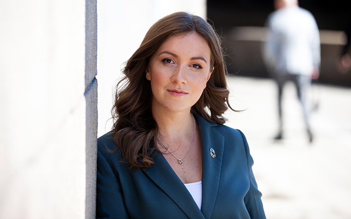 Kvinna med brunt hår i blå kavaj står vid en vägg, utomhus, med en oskarp bakgrund