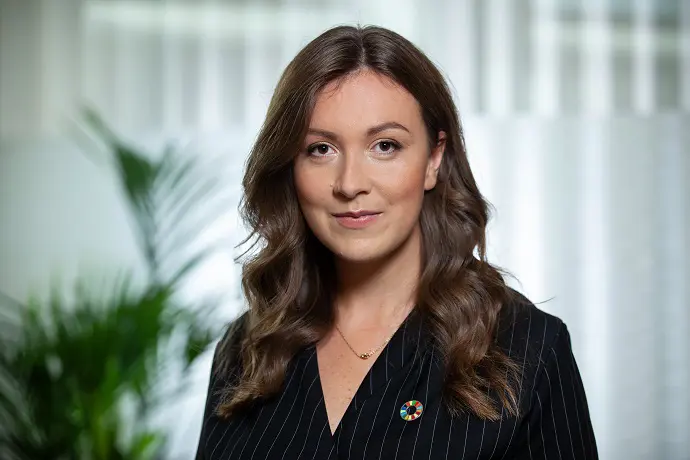 Kvinna i svart kavaj framför en suddig bakgrund med gröna växter och vita gardiner