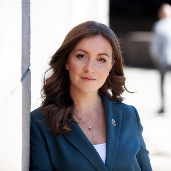 Kvinna i blå kavaj med brunt hår står utomhus mot en solbelyst vägg