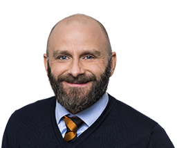 Man med skägg och rakat huvud, klädd i mörkblå tröja och randig slips, ler mot kameran