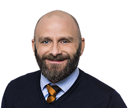 Man med skägg och rakat huvud, klädd i mörkblå tröja och randig slips, ler mot kameran