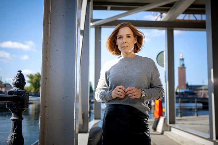 Kvinna i grå tröja står vid vattnet med Stadshuset i bakgrunden under en solig dag i Stockholm