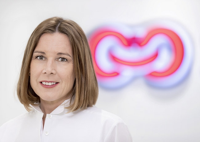 Kvinna med axellångt brunt hår och vit blus står framför en suddig, färgglad logotyp