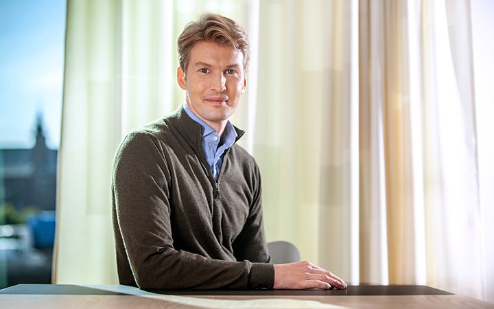 Man i grå tröja sitter vid ett bord framför ett fönster med ljusgardiner