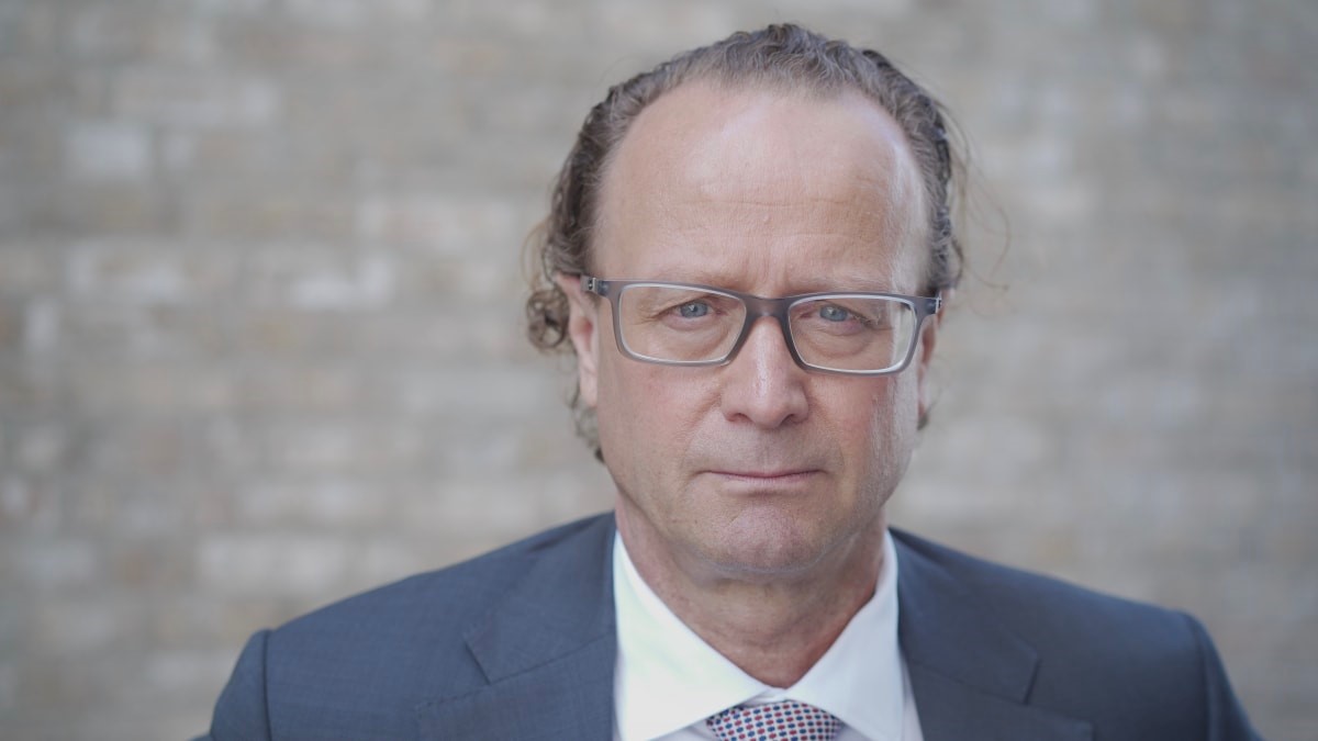 Man i kostym och glasögon mot en tegelvägg, i en formell och allvarlig pose