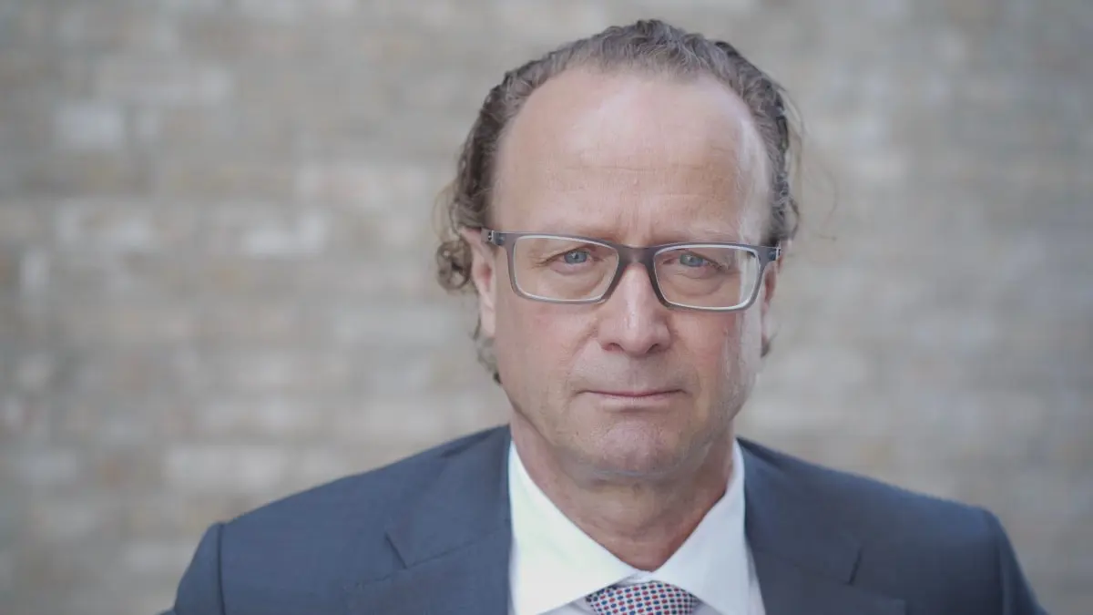 Man i kostym och glasögon mot en tegelvägg, i en formell och allvarlig pose