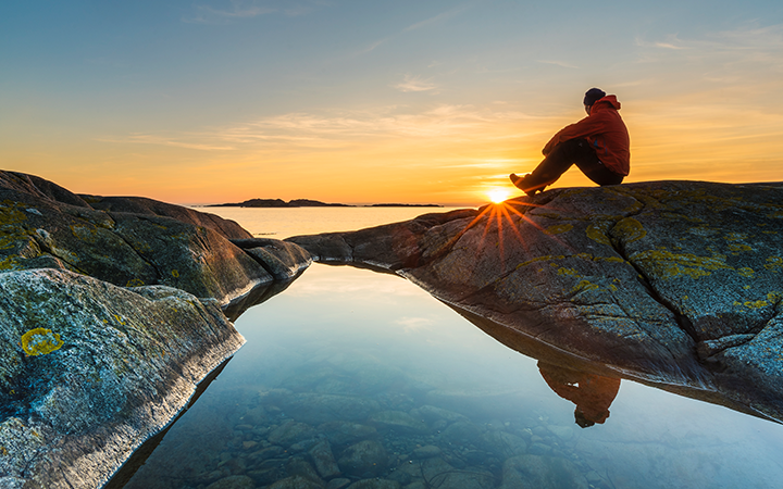 Person sitter på klippa vid solnedgång vid havet, med klart vatten och reflektioner. Avkoppling i skärgårdsmiljö