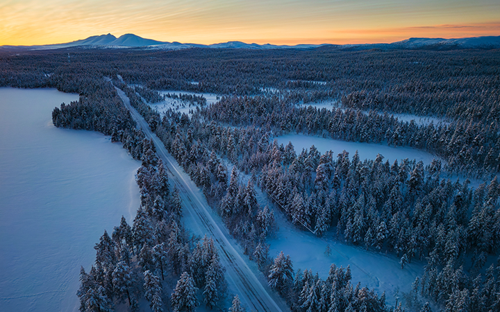 Vinterlandskap i norra Sverige med snötäckta träd, en lång väg och avlägsna berg vid solnedgången
