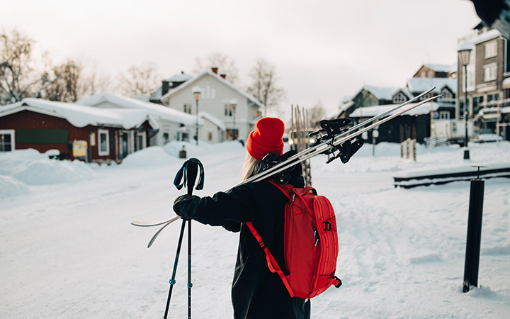 Person klädd i vinterkläder med röd ryggsäck bär skidor genom en snöig by