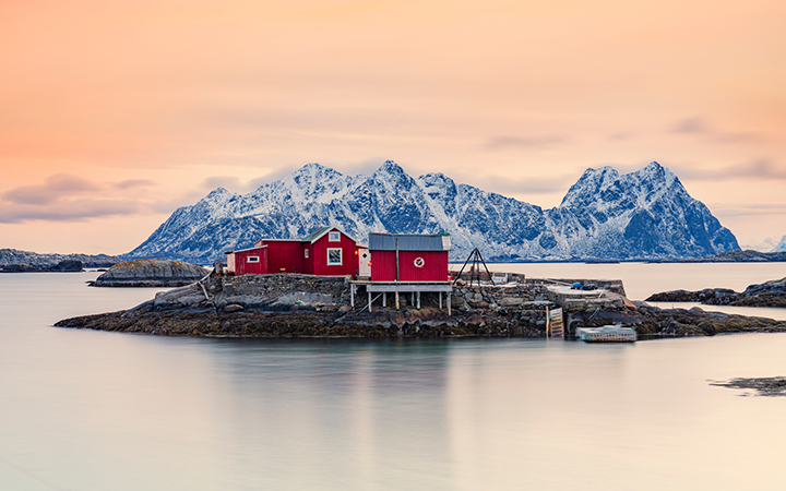 Röd stuga på en liten ö vid Lofoten med snötäckta berg i bakgrunden och en pastellfärgad himmel