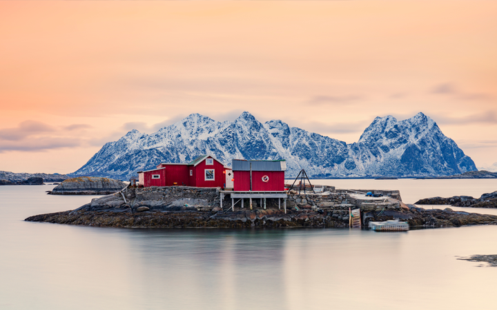 Röda stugor vid havet med snötäckta berg i bakgrunden under en solnedgång i Lofoten, Norge