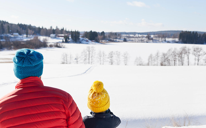 Två personer med färgglada mössor blickar ut över ett snötäckt landskap i Sverige, med skog och hus i bakgrunden