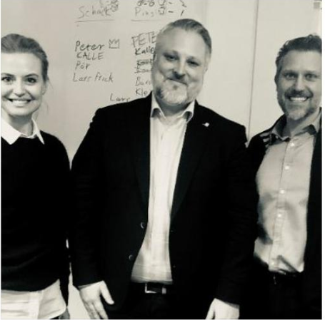 Tre personer i kontorskläder står framför en whiteboard med text och namn