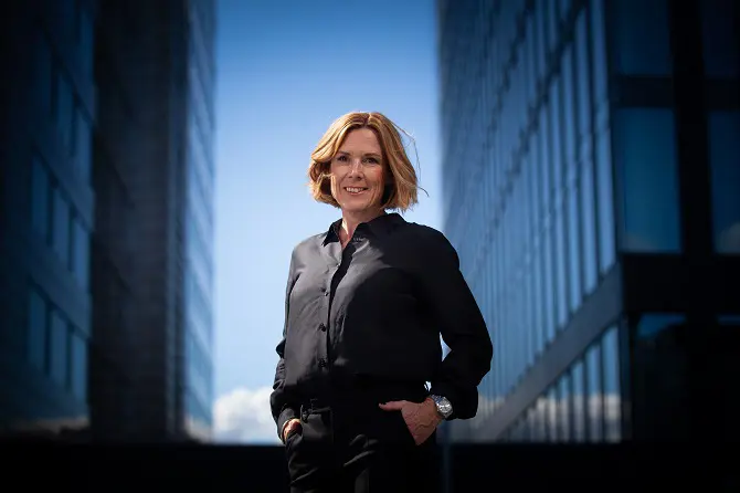 Kvinna i svart skjorta står mellan två höga byggnader, ler mot kameran under en klarblå himmel