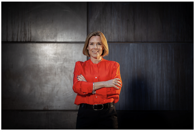 Kvinna i röd skjorta med armarna i kors framför en mörk bakgrund