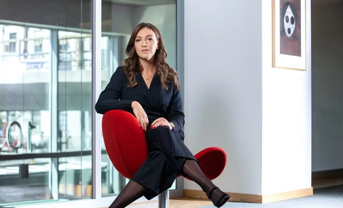 Kvinna i elegant mörk kostym sitter på röd designstol i modernt kontorslandskap