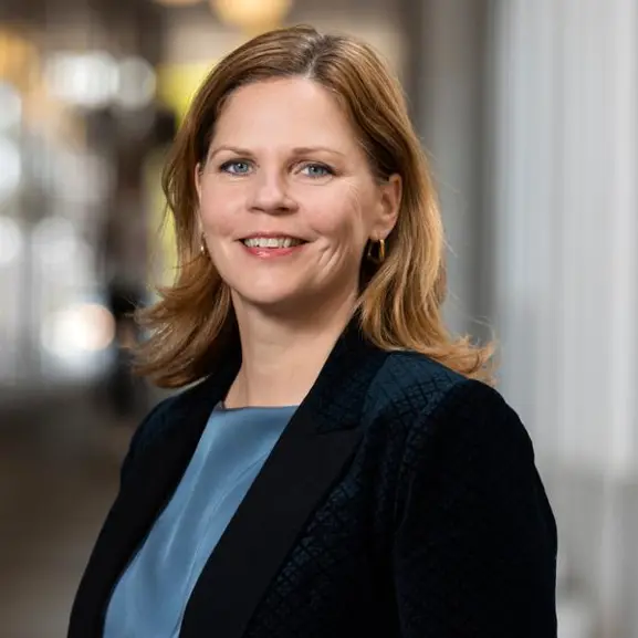 Kvinna med blont hår och blå kavaj ler i en kontorsmiljö