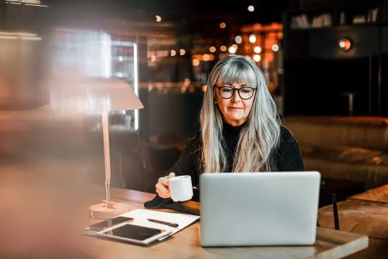 Äldre kvinna med långt grått hår och glasögon arbetar på en bärbar dator och dricker kaffe i ett modernt kafé