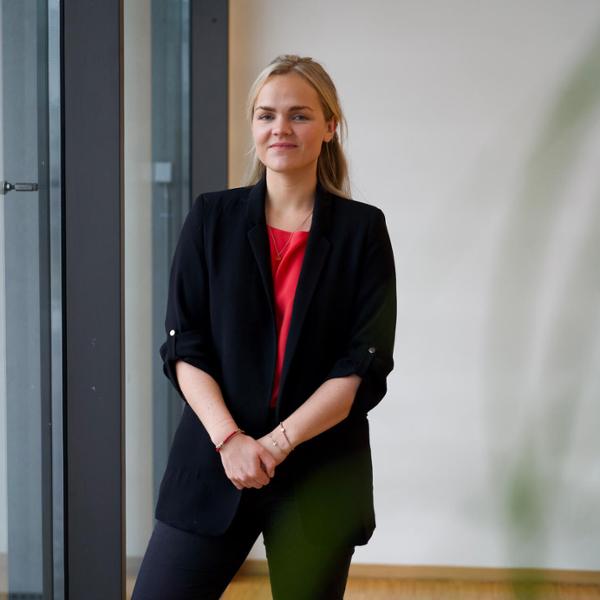 En kvinna i svart kavaj och röd topp står i ett modernt kontorsmiljö med fönster i bakgrunden