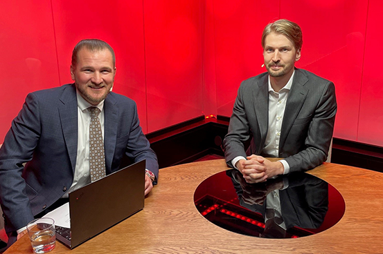 Två män i kostymer sitter vid ett runt bord med en dator i ett modernt rum med röd bakgrund