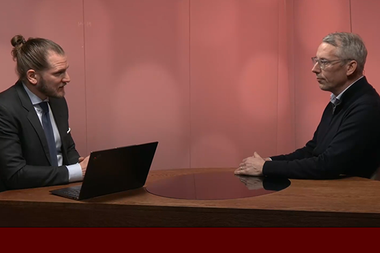 Två män i formellt samtal vid ett bord, en med laptop, i en rum med rosa belysning