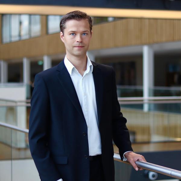 Man i mörk kostym står i modernt kontorslandskap, lutar sig mot räcke