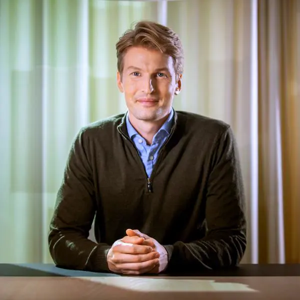 Man i mörk tröja och ljusblå skjorta sitter vid bord, omgiven av ljusa gardiner