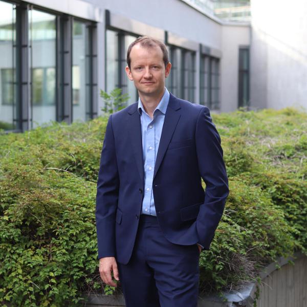 Man i mörkblå kostym står utomhus framför en modern byggnad med glasfönster, omgiven av grönska