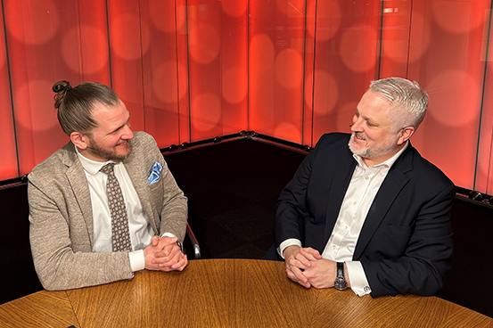 Två män som diskuterar vid ett runt bord, omgivna av röda bakgrundsskärmar. Ena mannen bär en beige kavaj och slips, den andra mannen bär en mörk kostym
