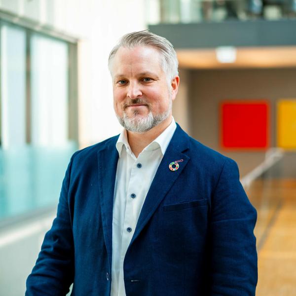 Man i mörkblå kavaj och vit skjorta står inomhus med färgglada tavlor i bakgrunden