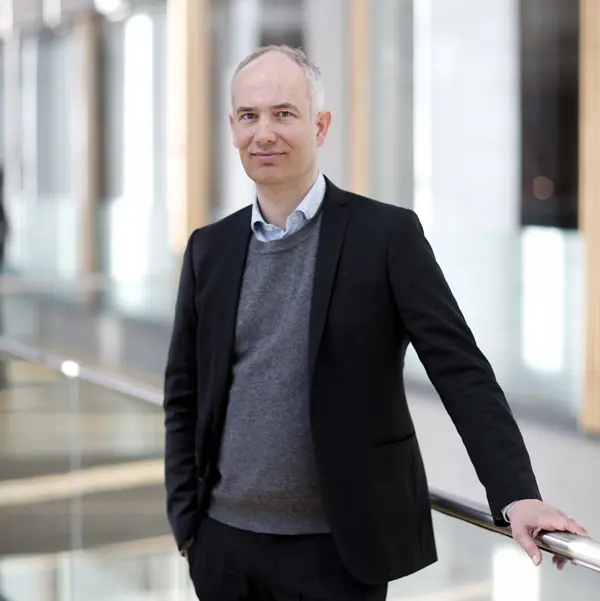 En man i kostym står vid ett modernt glasräcke inomhus, ser in i kameran