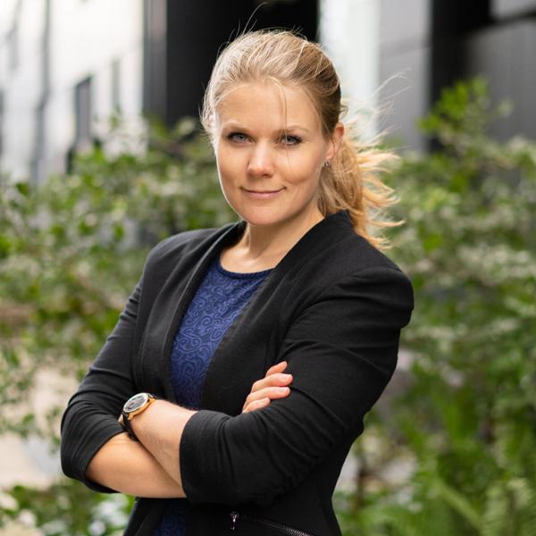 Kvinna i affärskläder utomhus med armarna i kors, bakgrund av gröna växter och moderna byggnader