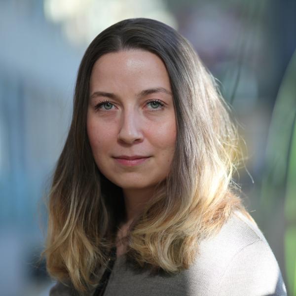 Kvinna med långt brunt hår och naturlig sminkning tittar in i kameran, suddig bakgrund med dämpade färger