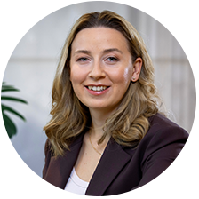 En leende kvinna med blont hår i en brun kavaj, sittande framför en ljus bakgrund med en grön växt