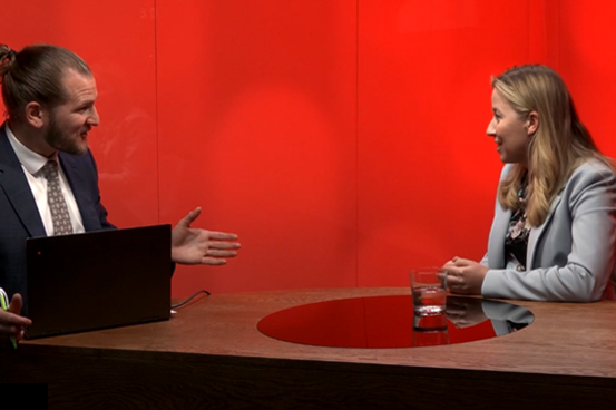 Två personer diskuterar vid ett bord med en laptop på, mot en röd bakgrund